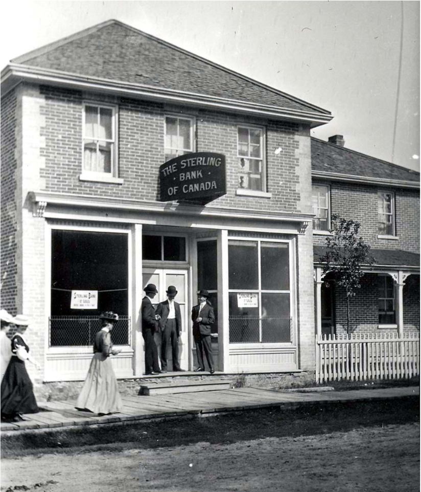 Two story brick building with 3 men standing at the front door. Three ladies in long dresses passing by along the wooden sidewalk.